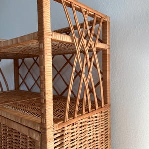 May include: A tall, natural wicker shelf unit with a woven design. The shelf has three shelves and a cabinet door. The cabinet door is closed.