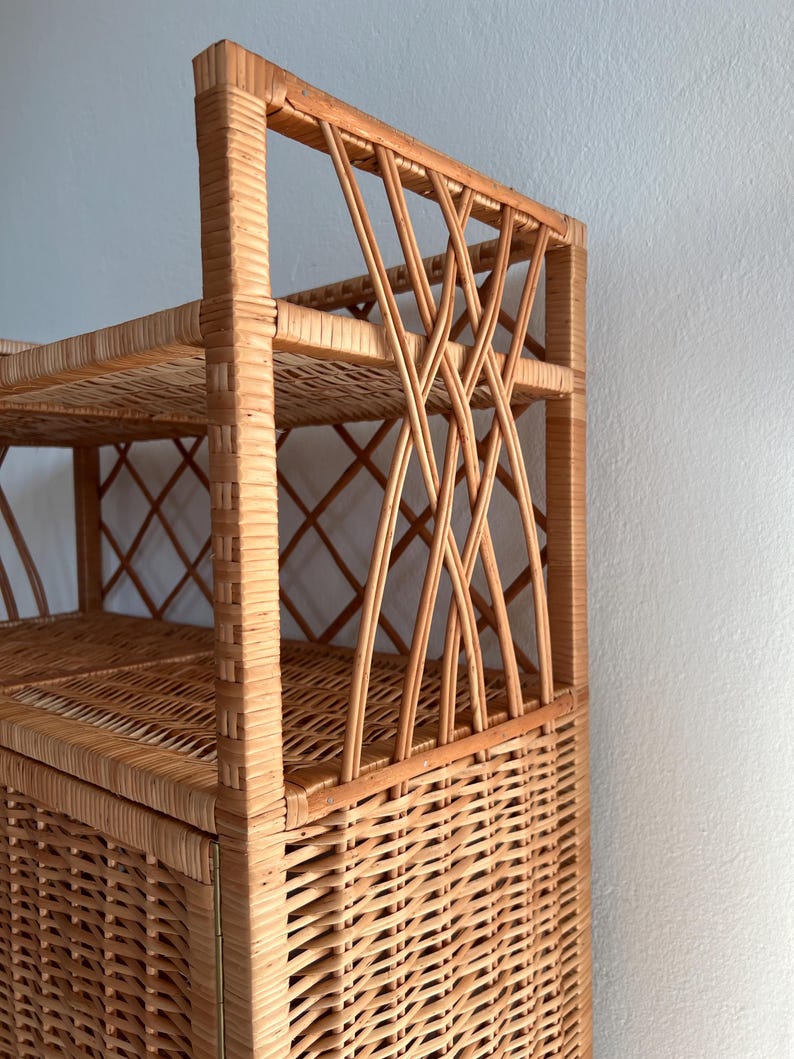 May include: A tall, natural wicker shelf unit with a woven design. The shelf has three shelves and a cabinet door. The cabinet door is closed.