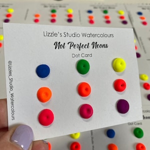 May include: A white card with the text "Lizzie's Studio Watercolours Not Perfect Neons Dot Card" displays a selection of neon paint dots in various colours. The dots include blue, orange, pink, green, yellow, and purple. Other cards with similar dots are visible.