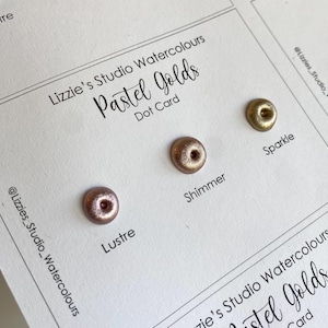 May include: A card with four small dots of metallic paint in shades of gold. The card is labelled "Lizzie's Studio Watercolours Pastel Golds Dot Card" and each dot is labelled with a name: "Lustre", "Shimmer", "Sparkle", and "Gold".