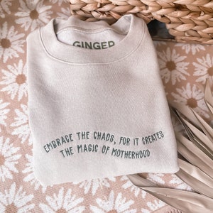 May include: A light beige sweatshirt with the text "EMBRACE THE CHAOS, FOR IT CREATES THE MAGIC OF MOTHERHOOD" embroidered in green on the front. The sweatshirt has the word "GINGED" printed in green on the neckline.
