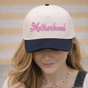 May include: A baseball cap with a cream-colored crown and a navy blue brim. The word "Motherhood" is embroidered in pink script across the front. The cap is worn by a person wearing denim overalls.