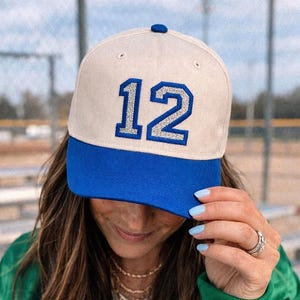 Puede incluir: Una gorra de b&eacute;isbol con una corona de color crema y una visera azul vibrante. La parte delantera de la gorra muestra el n&uacute;mero 12 en una fuente llamativa con borde azul y relleno de purpurina plateada. Incluye un peque&ntilde;o bot&oacute;n azul arriba.