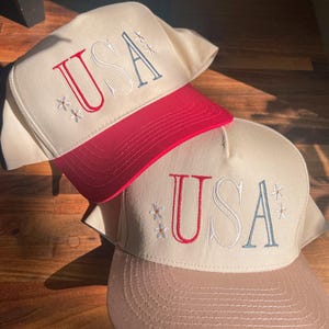 May include: Two baseball caps with the word "USA" embroidered on the front. One cap has a red brim and the other has a tan brim. Both caps have a cream-colored crown and small floral embroidery.