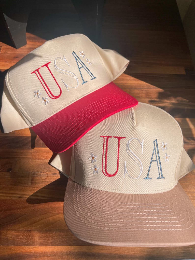 May include: Two baseball caps with the word "USA" embroidered on the front. One cap has a red brim and the other has a tan brim. Both caps have a cream-colored crown and small floral embroidery.