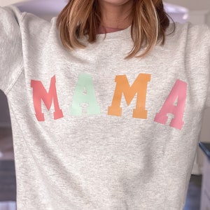 K&ouml;nnte beinhalten: Ein graues Sweatshirt mit dem Wort "MAMA" in Regenbogenfarben bedruckt.