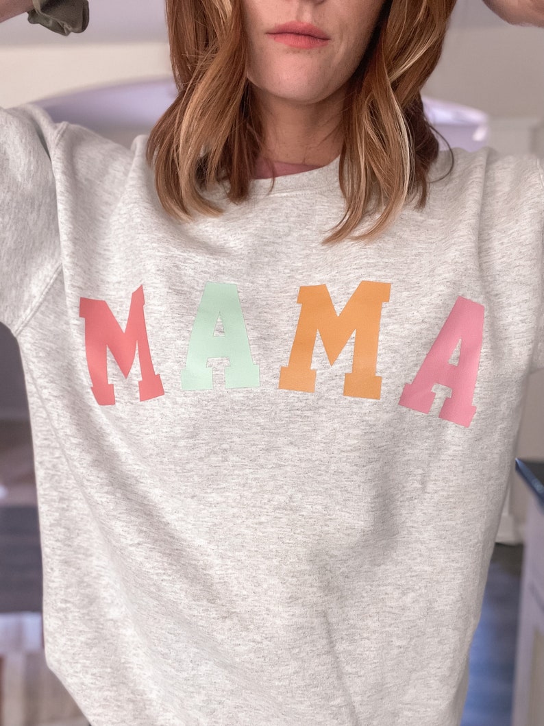 K&ouml;nnte beinhalten: Ein graues Sweatshirt mit dem Wort "MAMA" in Regenbogenfarben bedruckt.