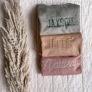 Puede incluir: Tres sudaderas plegadas con nombres personalizados: Jaxson, Lettie y Madison. Las sudaderas son en tonos de verde, beige y rosa.