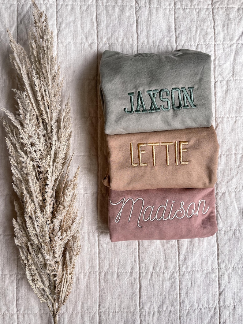 Puede incluir: Tres sudaderas plegadas con nombres personalizados: Jaxson, Lettie y Madison. Las sudaderas son en tonos de verde, beige y rosa.