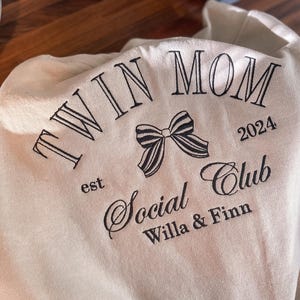 May include: Beige sweatshirt with black embroidery that reads "Twin Mom est 2024 Social Club Willa & Finn" with a black and white bow.