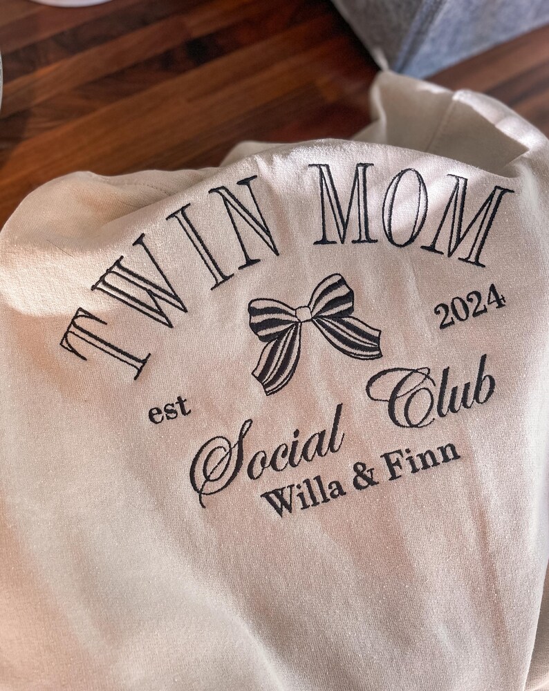 May include: Beige sweatshirt with black embroidery that reads "Twin Mom est 2024 Social Club Willa & Finn" with a black and white bow.