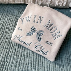 May include: A beige sweatshirt with the text "Twin Mom Social Club Est. 2025" embroidered in blue thread. A blue bow is embroidered in the center of the text.