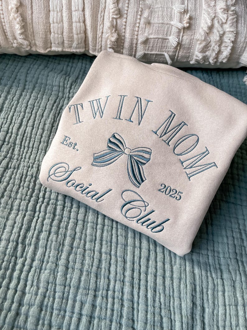 May include: A beige sweatshirt with the text "Twin Mom Social Club Est. 2025" embroidered in blue thread. A blue bow is embroidered in the center of the text.