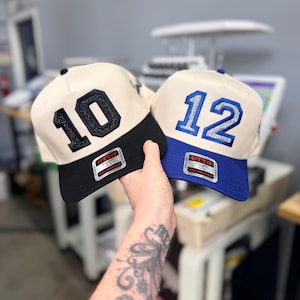 Puede incluir: Dos gorras de b&eacute;isbol con coronas de color crema y viseras estructuradas. La gorra de la izquierda tiene una visera negra y el n&uacute;mero 10 en purpurina negra. La de la derecha tiene una visera azul y el n&uacute;mero 12 en purpurina plateada. Ambas tienen etiquetas.