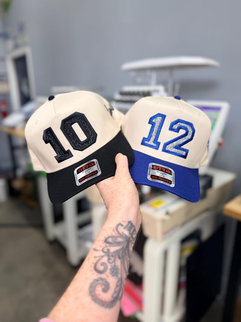 Puede incluir: Dos gorras de b&eacute;isbol con coronas de color crema y viseras estructuradas. La gorra de la izquierda tiene una visera negra y el n&uacute;mero 10 en purpurina negra. La de la derecha tiene una visera azul y el n&uacute;mero 12 en purpurina plateada. Ambas tienen etiquetas.
