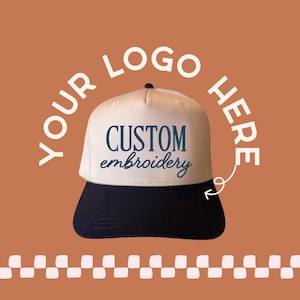 May include: A two-tone baseball cap with a cream-colored front panel and a dark navy blue brim. The front panel features the dark navy blue text "CUSTOM embroidery" in a combination of block and script fonts. An arrow points to the cap.