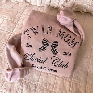 May include: A pink sweatshirt with the text "Twin Mom Social Club" embroidered on the front. The text is in black and includes a black bow and the year "2024". The text "Est." and "David & Dean" are also embroidered on the sweatshirt.