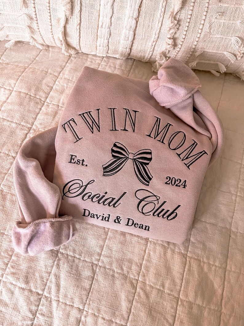 May include: A pink sweatshirt with the text "Twin Mom Social Club" embroidered on the front. The text is in black and includes a black bow and the year "2024". The text "Est." and "David & Dean" are also embroidered on the sweatshirt.
