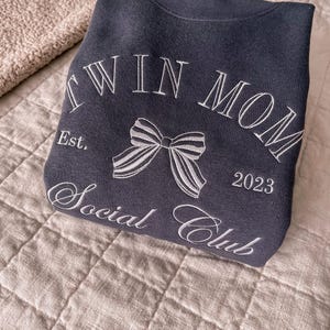 May include: A dark gray sweatshirt with the text "Twin Mom Social Club Est. 2023" and a white bow embroidered on the front.