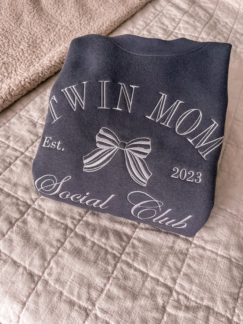 May include: A dark gray sweatshirt with the text "Twin Mom Social Club Est. 2023" and a white bow embroidered on the front.