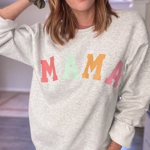 K&ouml;nnte beinhalten: Ein grauer Pullover mit dem Wort "MAMA" in Regenbogenfarben bedruckt. Die Buchstaben sind in einer fetten, blockartigen Schrift.