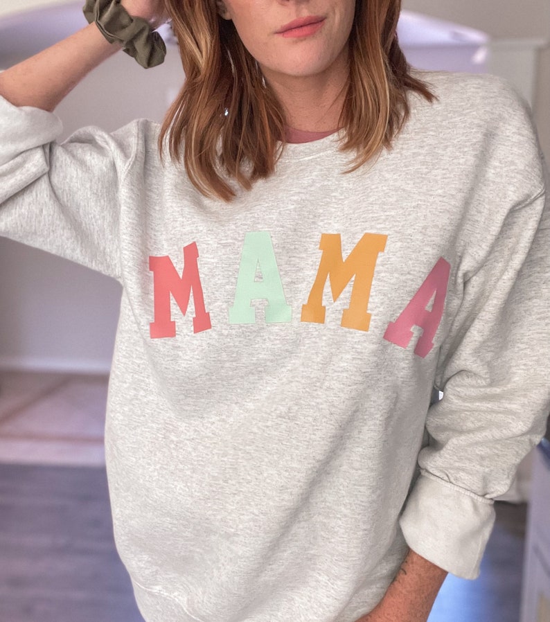 K&ouml;nnte beinhalten: Ein grauer Pullover mit dem Wort "MAMA" in Regenbogenfarben bedruckt. Die Buchstaben sind in einer fetten, blockartigen Schrift.
