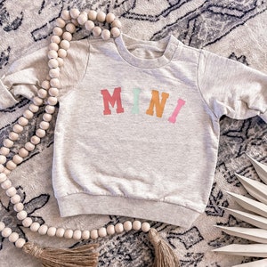 K&ouml;nnte beinhalten: Ein hellgrauer Pullover mit dem Wort "MINI" in bunten Buchstaben auf der Vorderseite. Der Pullover liegt auf einem Teppich mit einer Holzperlenkette.