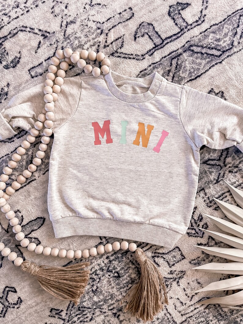 K&ouml;nnte beinhalten: Ein hellgrauer Pullover mit dem Wort "MINI" in bunten Buchstaben auf der Vorderseite. Der Pullover liegt auf einem Teppich mit einer Holzperlenkette.