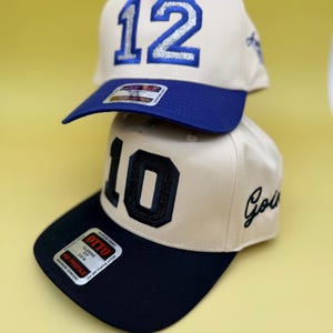 Puede incluir: Dos gorras de b&eacute;isbol de color beige apiladas verticalmente. La gorra superior presenta el n&uacute;mero 12 en purpurina azul con visera azul. La gorra inferior presenta el n&uacute;mero 10 en purpurina negra con visera negra y la palabra Gold bordada.