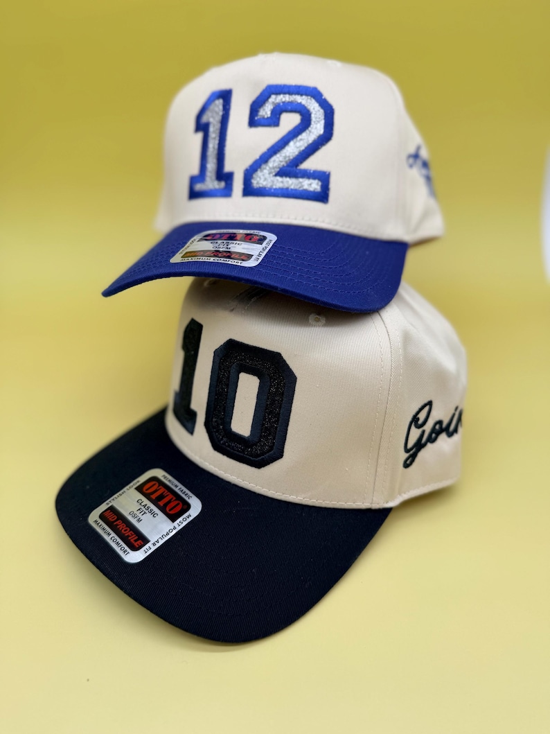 Puede incluir: Dos gorras de b&eacute;isbol de color beige apiladas verticalmente. La gorra superior presenta el n&uacute;mero 12 en purpurina azul con visera azul. La gorra inferior presenta el n&uacute;mero 10 en purpurina negra con visera negra y la palabra Gold bordada.