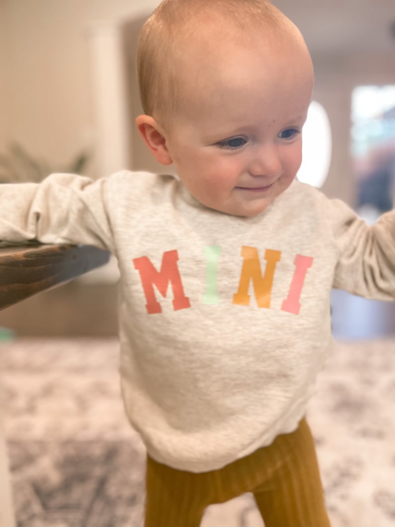 K&ouml;nnte beinhalten: Ein Kleinkind tr&auml;gt einen hellgrauen Pullover mit dem Wort "MINI" in bunten Buchstaben bedruckt. Das Kind tr&auml;gt eine braune Cordhose.