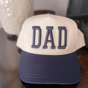 Puede incluir: Una gorra de b&eacute;isbol blanca y azul marino con la palabra "DAD" bordada en azul marino en la parte delantera.