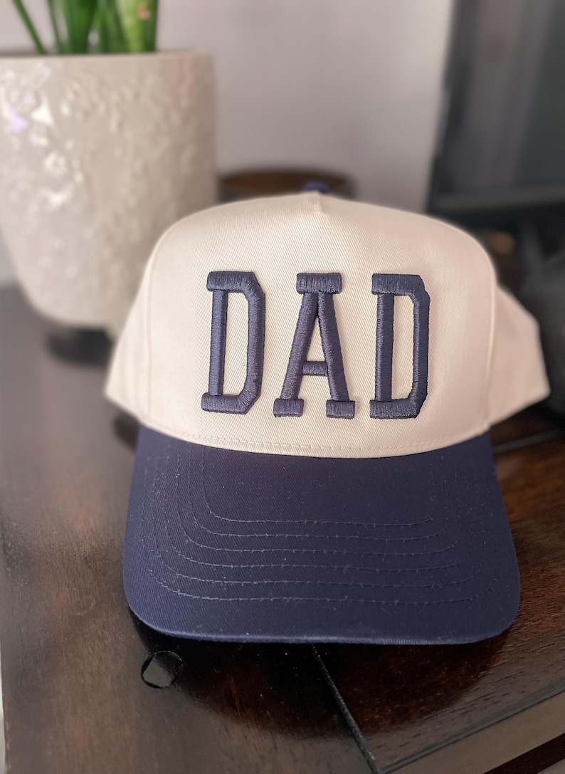 Puede incluir: Una gorra de b&eacute;isbol blanca y azul marino con la palabra "DAD" bordada en azul marino en la parte delantera.