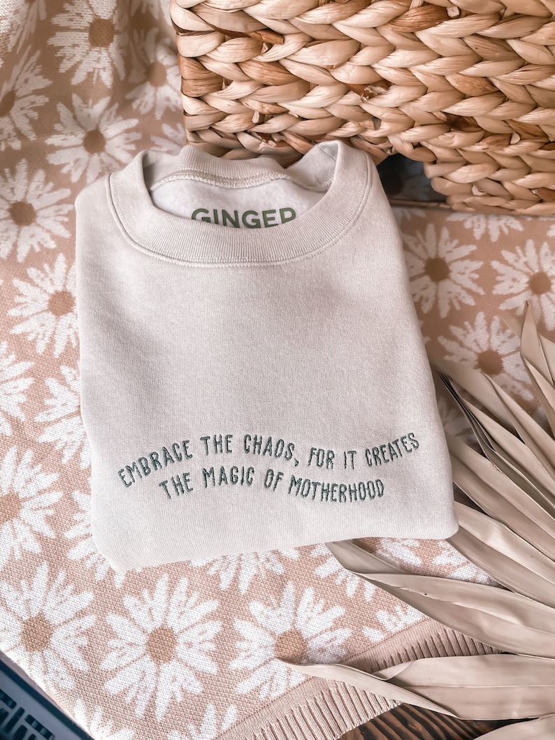 May include: A light beige sweatshirt with the text "EMBRACE THE CHAOS, FOR IT CREATES THE MAGIC OF MOTHERHOOD" embroidered in green on the front. The sweatshirt has the word "GINGED" printed in green on the neckline.