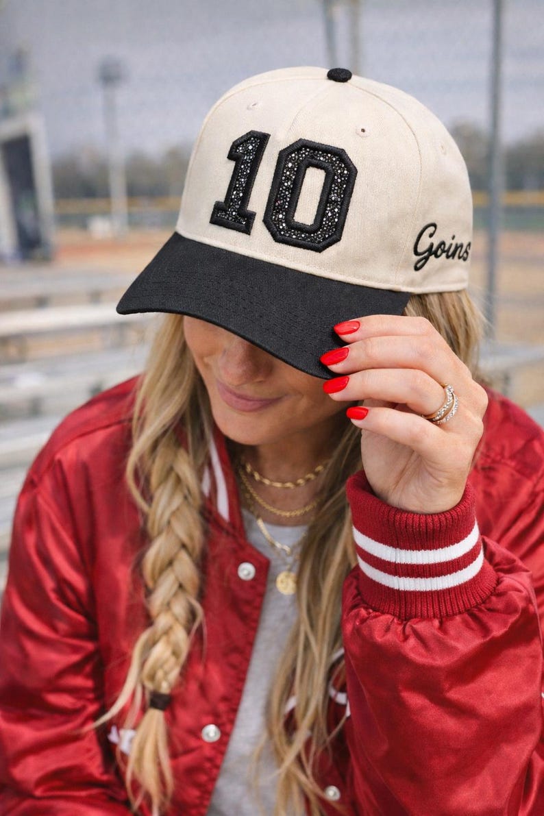 May include: A beige baseball cap with a black brim, featuring the number "10" embellished with rhinestones and the word "Goins" embroidered on the side. The cap is worn with a red satin jacket.