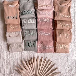 Puede incluir: Una pila de 10 sudaderas beige, rosa y gris con diferentes nombres bordados en cada una. Los nombres son: Ginger, Dawson, Axel, Jaxson, Ava, Patrick, Grace, Nora, Lathan, Cole, Harper, Lettie, Raegan, Dax, Madison y Audriella.