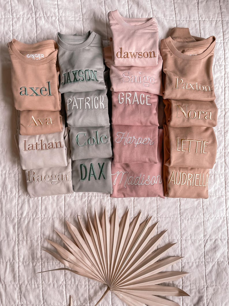 Puede incluir: Una pila de 10 sudaderas beige, rosa y gris con diferentes nombres bordados en cada una. Los nombres son: Ginger, Dawson, Axel, Jaxson, Ava, Patrick, Grace, Nora, Lathan, Cole, Harper, Lettie, Raegan, Dax, Madison y Audriella.