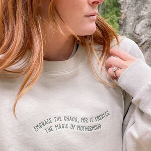 May include: A light beige sweatshirt with the text "Embrace the chaos, for it creates the magic of motherhood" embroidered in dark gray.