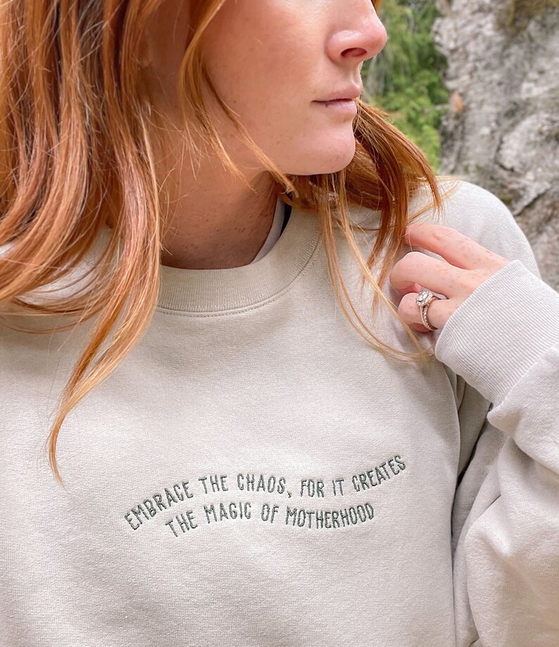 May include: A light beige sweatshirt with the text "Embrace the chaos, for it creates the magic of motherhood" embroidered in dark gray.