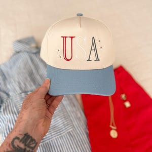 May include: A baseball cap with a cream-colored crown and a light blue brim. The word "USA" is embroidered on the front in red, white, and dark blue, with small star accents. The hat is a patriotic accessory.