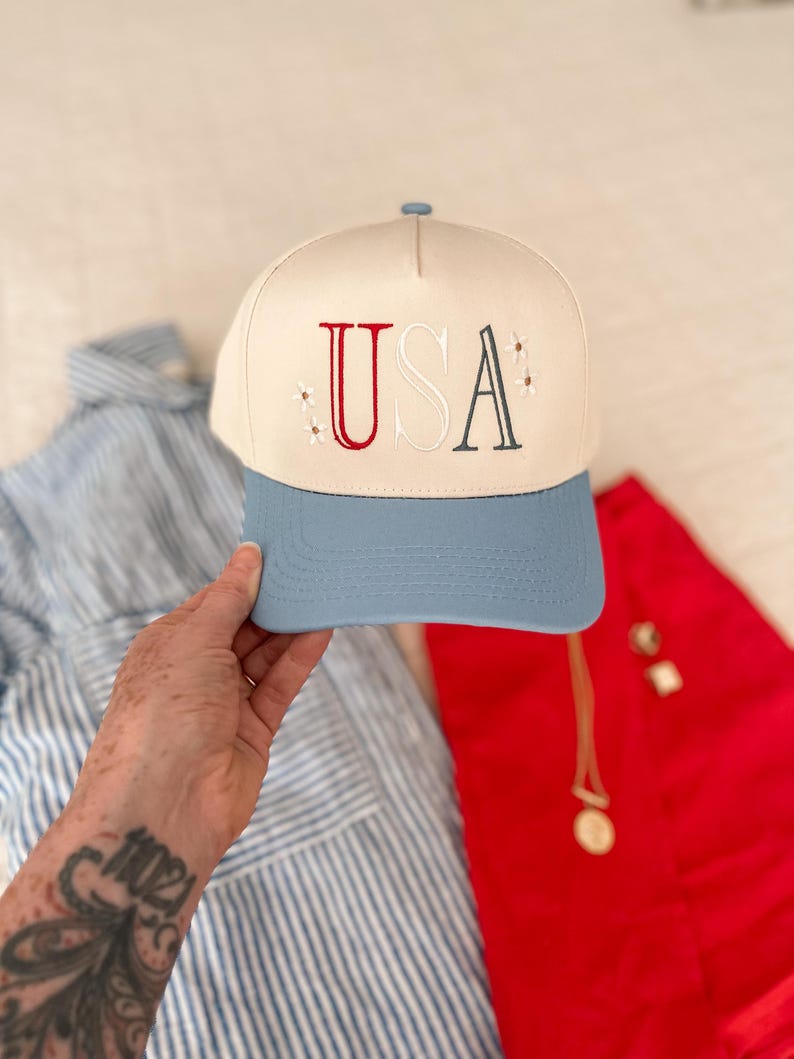 May include: A baseball cap with a cream-colored crown and a light blue brim. The word "USA" is embroidered on the front in red, white, and dark blue, with small star accents. The hat is a patriotic accessory.