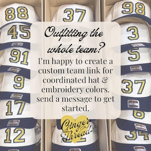 Puede incluir: Varias gorras de b&eacute;isbol blancas y azul marino dispuestas en cajas de cart&oacute;n. Cada gorra presenta un n&uacute;mero grande de color dorado brillante bordado en la parte delantera. El texto superpuesto dice: Outfitting the whole team? I'm happy to create a custom team link for coordinated hat & embroidery colors. send a message to get started. Ginger Thread.