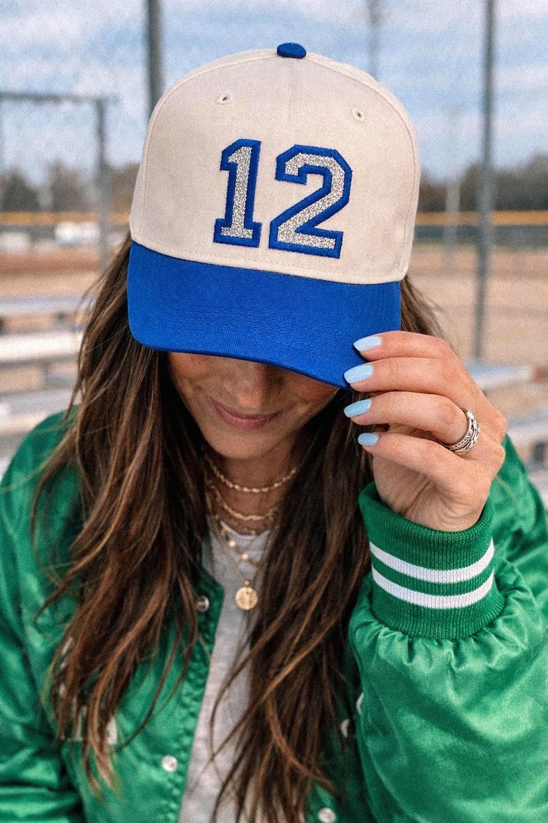 Puede incluir: Una gorra de b&eacute;isbol con una corona de color crema y una visera azul vibrante. La parte delantera de la gorra muestra el n&uacute;mero 12 en una fuente llamativa con borde azul y relleno de purpurina plateada. Incluye un peque&ntilde;o bot&oacute;n azul arriba.
