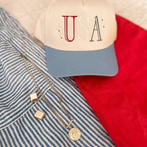 May include: A cream-colored baseball cap with "USA" embroidered in red and gray, accented with small star details. A gold necklace with a coin pendant and square earrings rest on a blue and white striped fabric, next to a red cloth.