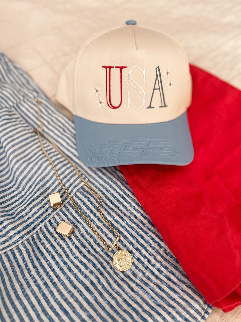 May include: A cream-colored baseball cap with "USA" embroidered in red and gray, accented with small star details. A gold necklace with a coin pendant and square earrings rest on a blue and white striped fabric, next to a red cloth.