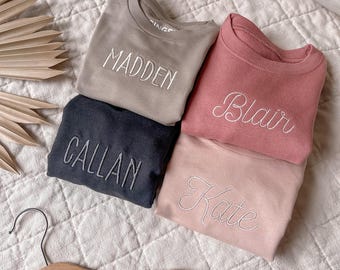 May include: Four personalized sweatshirts with different names embroidered on them: Madden, Blair, Callan, and Kate. The sweatshirts are in various colors, including beige, pink, and black. The sweatshirts are laid out on a white quilted fabric surface.