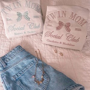 May include: Two beige sweatshirts with the text "Twin Mom Social Club" embroidered on them. The text is in a cursive font with a bow design above it. The sweatshirts have the years "2025" and "Est." embroidered on them. One sweatshirt has the names "Hayes & Parker" embroidered below the text and the other has the names "Charlotte & Madeline" embroidered below the text.
