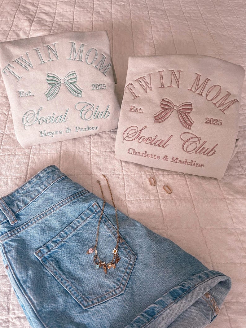 May include: Two beige sweatshirts with the text "Twin Mom Social Club" embroidered on them. The text is in a cursive font with a bow design above it. The sweatshirts have the years "2025" and "Est." embroidered on them. One sweatshirt has the names "Hayes & Parker" embroidered below the text and the other has the names "Charlotte & Madeline" embroidered below the text.