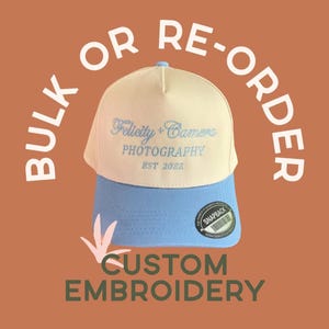 May include: A two-tone baseball cap with a cream-colored front panel and a light blue brim and back. The front features light blue embroidery reading Felicity + Camera PHOTOGRAPHY EST 2022. A circular black and white snapback sticker is on the brim.