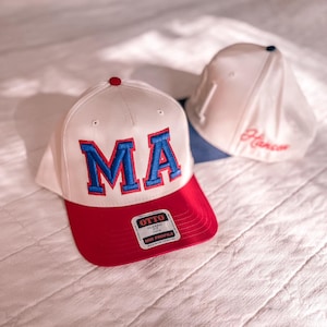 May include: Two baseball caps. One white cap with a red brim and the letters "MA" in blue. The other cap is white with a blue brim and the name "Hansen" in red. Both caps have a small button on top.